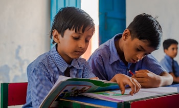 Two young boys wearing blue checkered school uniforms are sitting at a desk in a classroom, focused on their studies. One boy is reading from a colorful book while the other is writing diligently in a notebook. The classroom has a calm atmosphere with a blue door and simple, light-colored walls in the background.
