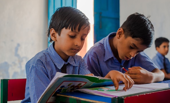 Two young boys wearing blue checkered school uniforms are sitting at a desk in a classroom, focused on their studies. One boy is reading from a colorful book while the other is writing diligently in a notebook. The classroom has a calm atmosphere with a blue door and simple, light-colored walls in the background.