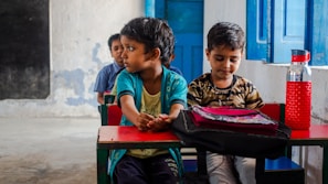 Children are sitting at small desks in a classroom setting. One child looks to the side while the others face forward. A red water bottle sits on the desk next to a pink and black bag. The classroom walls are painted blue and white, with a blue wooden door visible in the background.