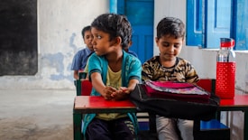 Children are sitting at small desks in a classroom setting. One child looks to the side while the others face forward. A red water bottle sits on the desk next to a pink and black bag. The classroom walls are painted blue and white, with a blue wooden door visible in the background.