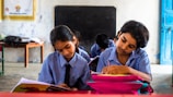 Two children in school uniforms are seated at desks, focused on writing in notebooks. The classroom has a chalkboard at the front, and a table with a backpack and other items in the background. The walls are lightly decorated, giving a simple yet structured environment.