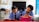 Two children in school uniforms are seated at desks, focused on writing in notebooks. The classroom has a chalkboard at the front, and a table with a backpack and other items in the background. The walls are lightly decorated, giving a simple yet structured environment.