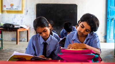 Two children in school uniforms are seated at desks, focused on writing in notebooks. The classroom has a chalkboard at the front, and a table with a backpack and other items in the background. The walls are lightly decorated, giving a simple yet structured environment.