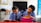 Two children in school uniforms are seated at desks, focused on writing in notebooks. The classroom has a chalkboard at the front, and a table with a backpack and other items in the background. The walls are lightly decorated, giving a simple yet structured environment.
