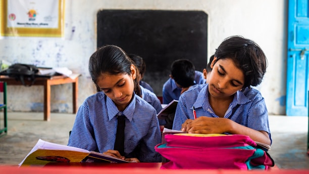 Two children in school uniforms are seated at desks, focused on writing in notebooks. The classroom has a chalkboard at the front, and a table with a backpack and other items in the background. The walls are lightly decorated, giving a simple yet structured environment.