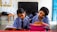 Two children in school uniforms are seated at desks, focused on writing in notebooks. The classroom has a chalkboard at the front, and a table with a backpack and other items in the background. The walls are lightly decorated, giving a simple yet structured environment.