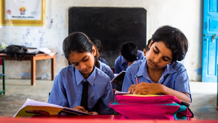Two children in school uniforms are seated at desks, focused on writing in notebooks. The classroom has a chalkboard at the front, and a table with a backpack and other items in the background. The walls are lightly decorated, giving a simple yet structured environment.