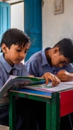 Two children in school uniforms are sitting at a desk in a classroom. They are focused on writing or drawing in their notebooks. The classroom setting includes open blue doors in the background, adding natural light to the scene.