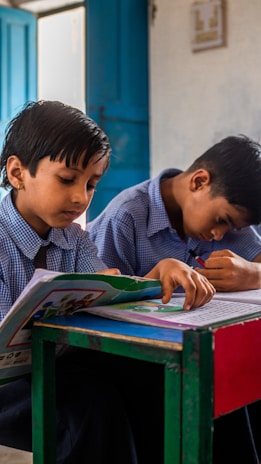 Two children in school uniforms are sitting at a desk in a classroom. They are focused on writing or drawing in their notebooks. The classroom setting includes open blue doors in the background, adding natural light to the scene.