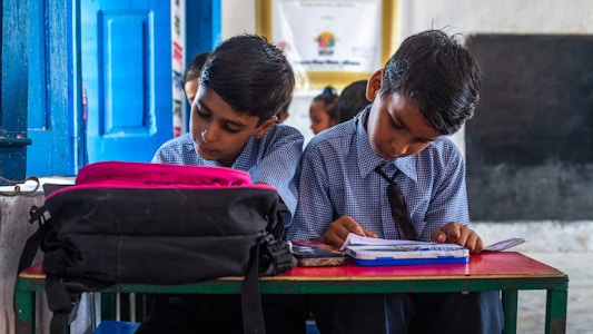 Two young boys in school uniforms are seated at a desk. They are focused on their studies, with books and a pencil case on the desk. A black backpack is placed nearby. The classroom setting includes a wooden bench, blue doors, and some educational posters on the wall.