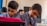 Two young boys are focused on their work, sitting side by side in a classroom setting. They wear matching blue checkered uniforms with ties, indicating a school environment. A blurred figure of another student is visible in the background. One of the boys has a bright pink item on his desk, possibly a backpack or book.