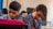Two young boys are focused on their work, sitting side by side in a classroom setting. They wear matching blue checkered uniforms with ties, indicating a school environment. A blurred figure of another student is visible in the background. One of the boys has a bright pink item on his desk, possibly a backpack or book.