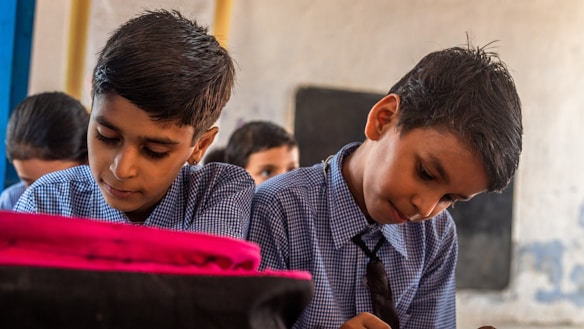 Two young boys are focused on their work, sitting side by side in a classroom setting. They wear matching blue checkered uniforms with ties, indicating a school environment. A blurred figure of another student is visible in the background. One of the boys has a bright pink item on his desk, possibly a backpack or book.