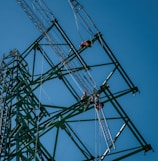 Several workers wearing safety gear are climbing a large metal lattice tower, which is possibly an electricity pylon. They use harnesses and ladders under a clear blue sky.
