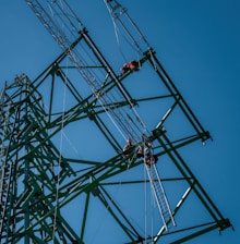 Several workers wearing safety gear are climbing a large metal lattice tower, which is possibly an electricity pylon. They use harnesses and ladders under a clear blue sky.
