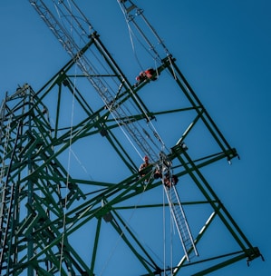 Several workers wearing safety gear are climbing a large metal lattice tower, which is possibly an electricity pylon. They use harnesses and ladders under a clear blue sky.