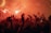 A crowd of people is gathered with arms raised, many holding up smartphones. Flares are visible, creating a bright, fiery red glow around the scene. The atmosphere is dense with excitement and energy, enhanced by the dramatic lighting.