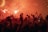 A crowd of people is gathered with arms raised, many holding up smartphones. Flares are visible, creating a bright, fiery red glow around the scene. The atmosphere is dense with excitement and energy, enhanced by the dramatic lighting.