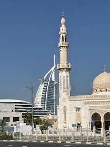 A modern skyscraper with a sail-like design stands prominently against a clear blue sky, positioned behind a traditional mosque featuring Islamic architectural elements such as a large domed roof and ornate minaret. The scene combines contemporary and traditional structures, surrounded by palm trees and set along a main road.