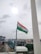 An Indian national flag is mounted on a pole, waving in the breeze against a cloudy sky. The background includes a cityscape with buildings and a few construction sites visible.