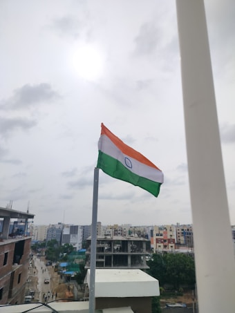 An Indian national flag is mounted on a pole, waving in the breeze against a cloudy sky. The background includes a cityscape with buildings and a few construction sites visible.
