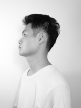 A black and white profile portrait of a young person wearing a plain white t-shirt. The background is a smooth, light grey, creating a stark contrast with the dark hair of the subject.