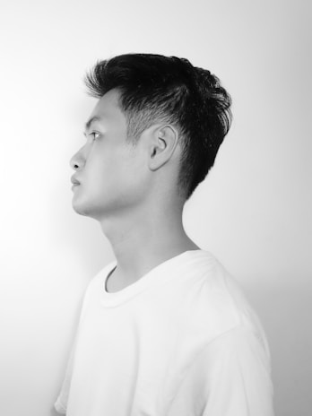A black and white profile portrait of a young person wearing a plain white t-shirt. The background is a smooth, light grey, creating a stark contrast with the dark hair of the subject.