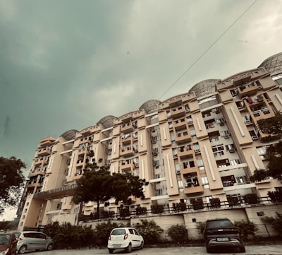 A multi-story residential building with a beige facade stands prominently under a cloudy sky. The architecture features multiple windows and balconies, some of which have drying clothes hanging. Below, several parked cars line the street in front of the building, and a few trees add greenery to the scene.