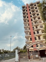 A tall multi-story residential building with multiple balconies, each with metal railings. The structure is painted primarily in beige and features a prominent red vertical accent. The entrance to the building has an ornate metal gate with a decorative archway. The sky is partly cloudy, and there is greenery around the property, including a tree on the right side of the image.