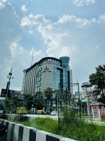 A large modern building with multiple floors and a curved structure is visible against a partly cloudy sky. There are palm trees and other greenery around the base of the building. Vehicles, including an auto rickshaw, are seen on the road in front of the building. A variety of signs and advertisements are present, and there is a fenced area of grass in the foreground.