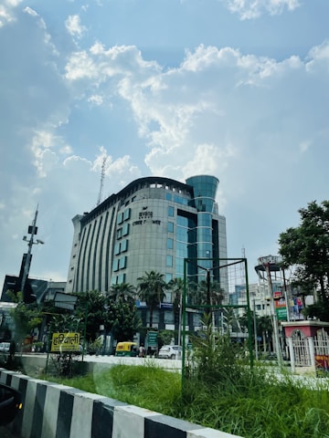 A large modern building with multiple floors and a curved structure is visible against a partly cloudy sky. There are palm trees and other greenery around the base of the building. Vehicles, including an auto rickshaw, are seen on the road in front of the building. A variety of signs and advertisements are present, and there is a fenced area of grass in the foreground.