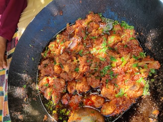 A large black cooking pan filled with a vibrant, spicy dish that appears to be curry, with chunks of meat and garnished with fresh green herbs. The dish has an oily surface that reflects light, and a metallic serving spoon is placed within the pan. Partial view of a person's hand and colorful fabric can be seen at the edge.