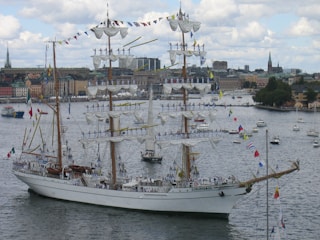 A large, elegant three-masted sailing ship adorned with numerous flags is navigating through a harbor. The masts are fully rigged with white sails, and multiple people in uniform are standing on the yardarms. In the background, a cityscape featuring older buildings and a few church spires is visible under a partly cloudy sky.