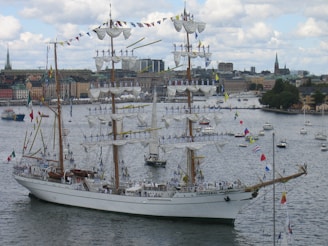 A large, elegant three-masted sailing ship adorned with numerous flags is navigating through a harbor. The masts are fully rigged with white sails, and multiple people in uniform are standing on the yardarms. In the background, a cityscape featuring older buildings and a few church spires is visible under a partly cloudy sky.