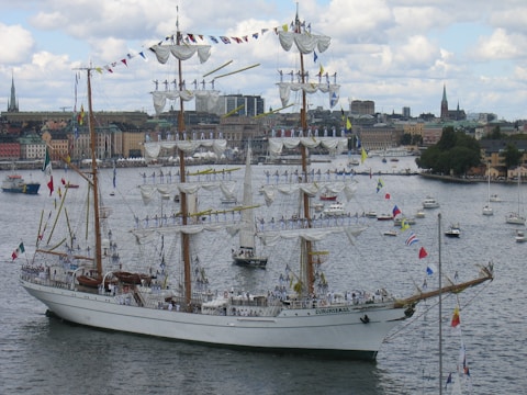 A large, elegant three-masted sailing ship adorned with numerous flags is navigating through a harbor. The masts are fully rigged with white sails, and multiple people in uniform are standing on the yardarms. In the background, a cityscape featuring older buildings and a few church spires is visible under a partly cloudy sky.