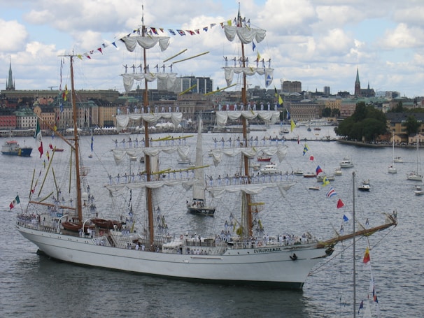 A large, elegant three-masted sailing ship adorned with numerous flags is navigating through a harbor. The masts are fully rigged with white sails, and multiple people in uniform are standing on the yardarms. In the background, a cityscape featuring older buildings and a few church spires is visible under a partly cloudy sky.