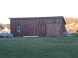 A rustic, weathered wooden barn with red paint stands in an open grassy area. A single window is visible on the left side, and double wooden doors occupy most of the front. To the left of the barn, there is a trailer or some kind of equipment. The background includes a small village or settlement with distant hills, under a clear sky.