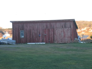 A rustic, weathered wooden barn with red paint stands in an open grassy area. A single window is visible on the left side, and double wooden doors occupy most of the front. To the left of the barn, there is a trailer or some kind of equipment. The background includes a small village or settlement with distant hills, under a clear sky.