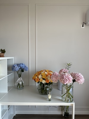 A stylish and organized interior space features a white desk with three vases of flowers. One vase holds blue hydrangeas, another is filled with a large bouquet of yellow and orange roses, and a third contains pink hydrangeas. A candle holder with a lit candle rests on the desk. The background is a minimalistic, light-colored wall with subtle paneling.