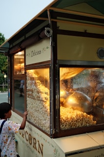 A young child stands by a popcorn cart, peering inside at the freshly popped popcorn. The cart is adorned with decorative text and illustrations, featuring warm lighting that highlights the popcorn machine's interior.
