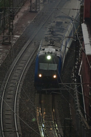 A train traveling on railway tracks, set against an industrial backdrop with overhead electrical wires. The tracks curve slightly as they move away from the vantage point, and the front of the train is illuminated by a bright headlight, casting reflections on the wet tracks.