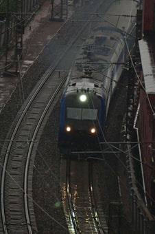 A train traveling on railway tracks, set against an industrial backdrop with overhead electrical wires. The tracks curve slightly as they move away from the vantage point, and the front of the train is illuminated by a bright headlight, casting reflections on the wet tracks.