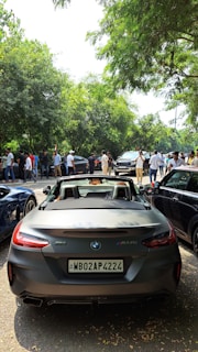 A convertible BMW Z4 with license plate WB02AP4224 is parked on a shaded street. Numerous people are gathered around various vehicles, under a canopy of lush green trees, suggesting a gathering or car meet.