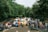 A group of people standing in front of a lineup of cars parked on a road, surrounded by lush green trees. The scene suggests a gathering or event, with attendees interacting casually and dressed in various casual outfits.