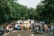 A group of people standing in front of a lineup of cars parked on a road, surrounded by lush green trees. The scene suggests a gathering or event, with attendees interacting casually and dressed in various casual outfits.