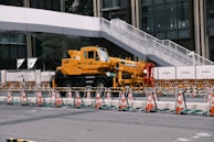 A yellow crane truck is parked in an urban construction site, surrounded by orange and white traffic cones and barricades. A pedestrian bridge arches overhead, and a modern building façade is visible in the background.