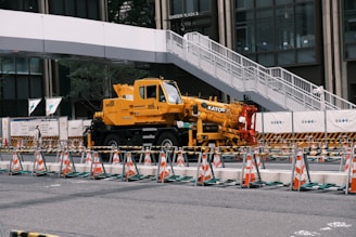 A yellow crane truck is parked in an urban construction site, surrounded by orange and white traffic cones and barricades. A pedestrian bridge arches overhead, and a modern building façade is visible in the background.