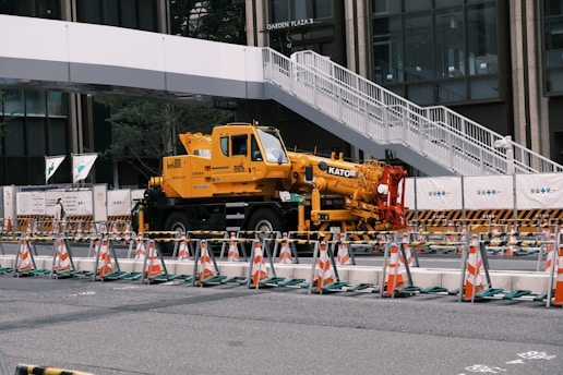 A yellow crane truck is parked in an urban construction site, surrounded by orange and white traffic cones and barricades. A pedestrian bridge arches overhead, and a modern building façade is visible in the background.