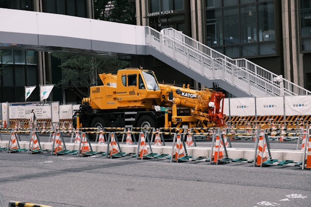 A yellow crane truck is parked in an urban construction site, surrounded by orange and white traffic cones and barricades. A pedestrian bridge arches overhead, and a modern building façade is visible in the background.
