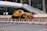 A yellow crane truck is parked in an urban construction site, surrounded by orange and white traffic cones and barricades. A pedestrian bridge arches overhead, and a modern building façade is visible in the background.